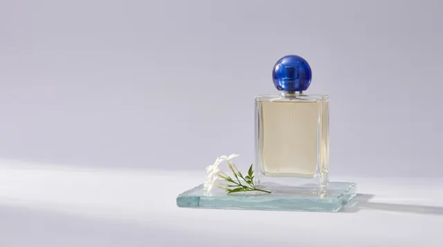 Wide perfume composition with a blue-capped glass bottle and jasmine on a translucent glass slab.